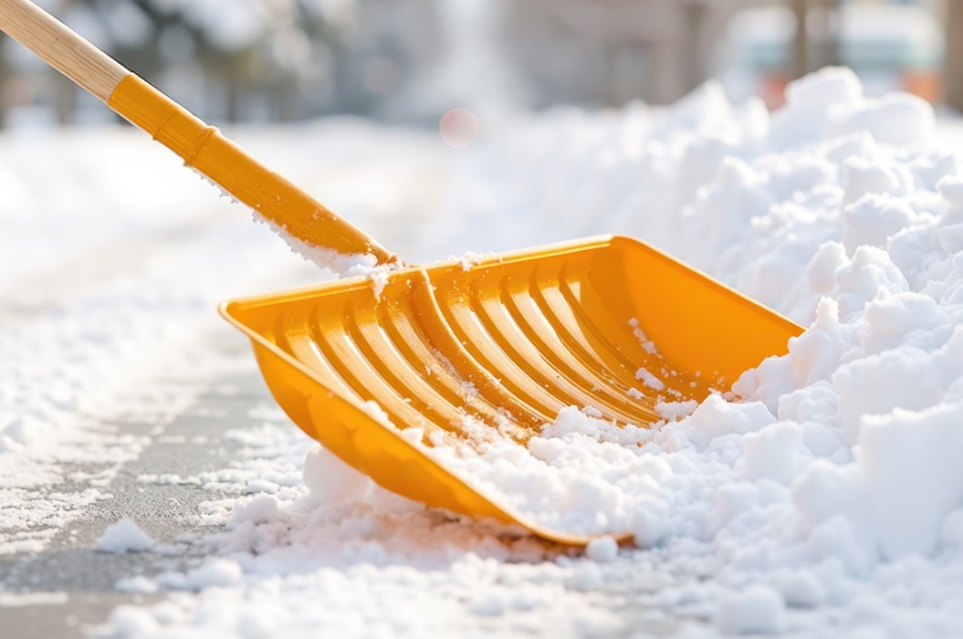 Nahaufnahme einer Person, die an einem kalten, strahlenden Wintermorgen eine gelbe Schneeschaufel schiebt, um einen verschneiten Gehweg zu räumen