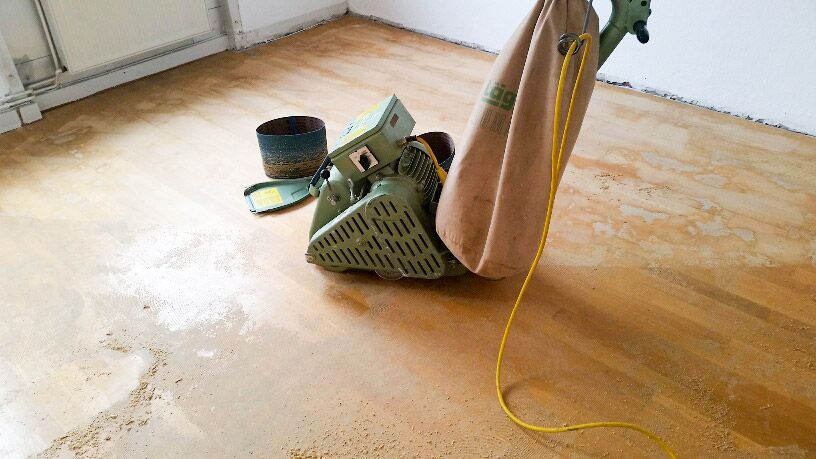 Eine gruene Bandschleifmaschine mit Staubbeutel steht auf einem Holzboden