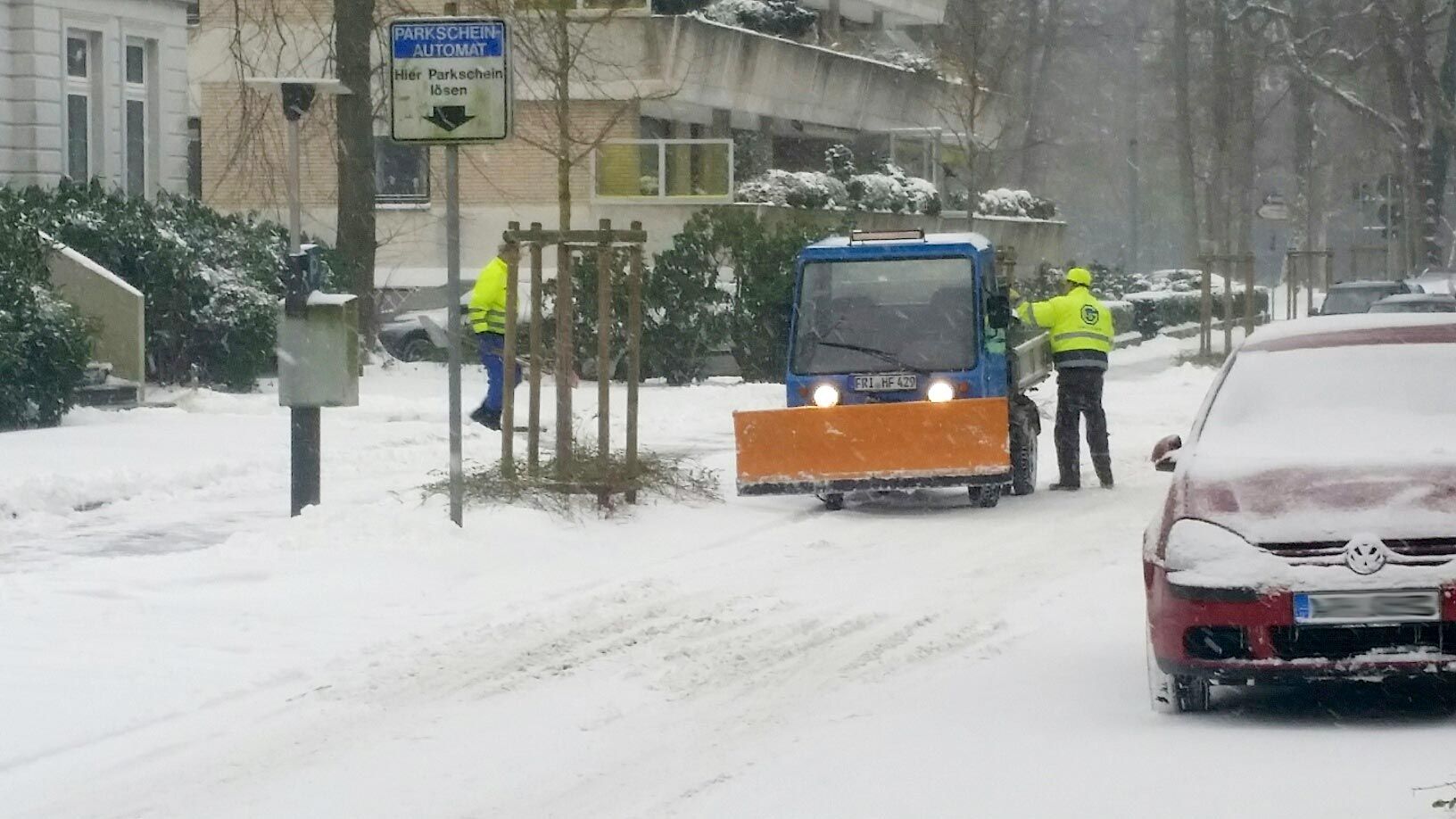 Zwei Arbeiter in gelben Warnwesten raeumen mit blauem Fahrzeug Schnee